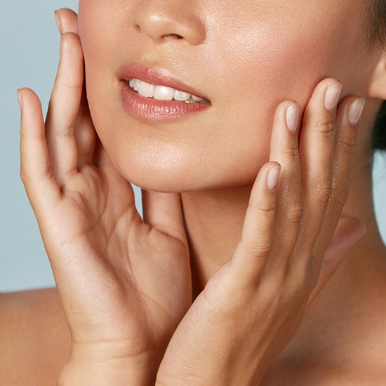 Zbliżenie na kobietę z nieskazitelną cerą, która delikatnie dotyka dłońmi policzków, prezentując promienny i zdrowy wygląd.