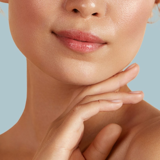 Zbliżenie na kobiecą twarz z delikatnym makijażem, gdzie usta są podkreślone błyszczącym, różowym odcieniem, a dłoń spoczywa pod brodą, akcentując gładką skórę.