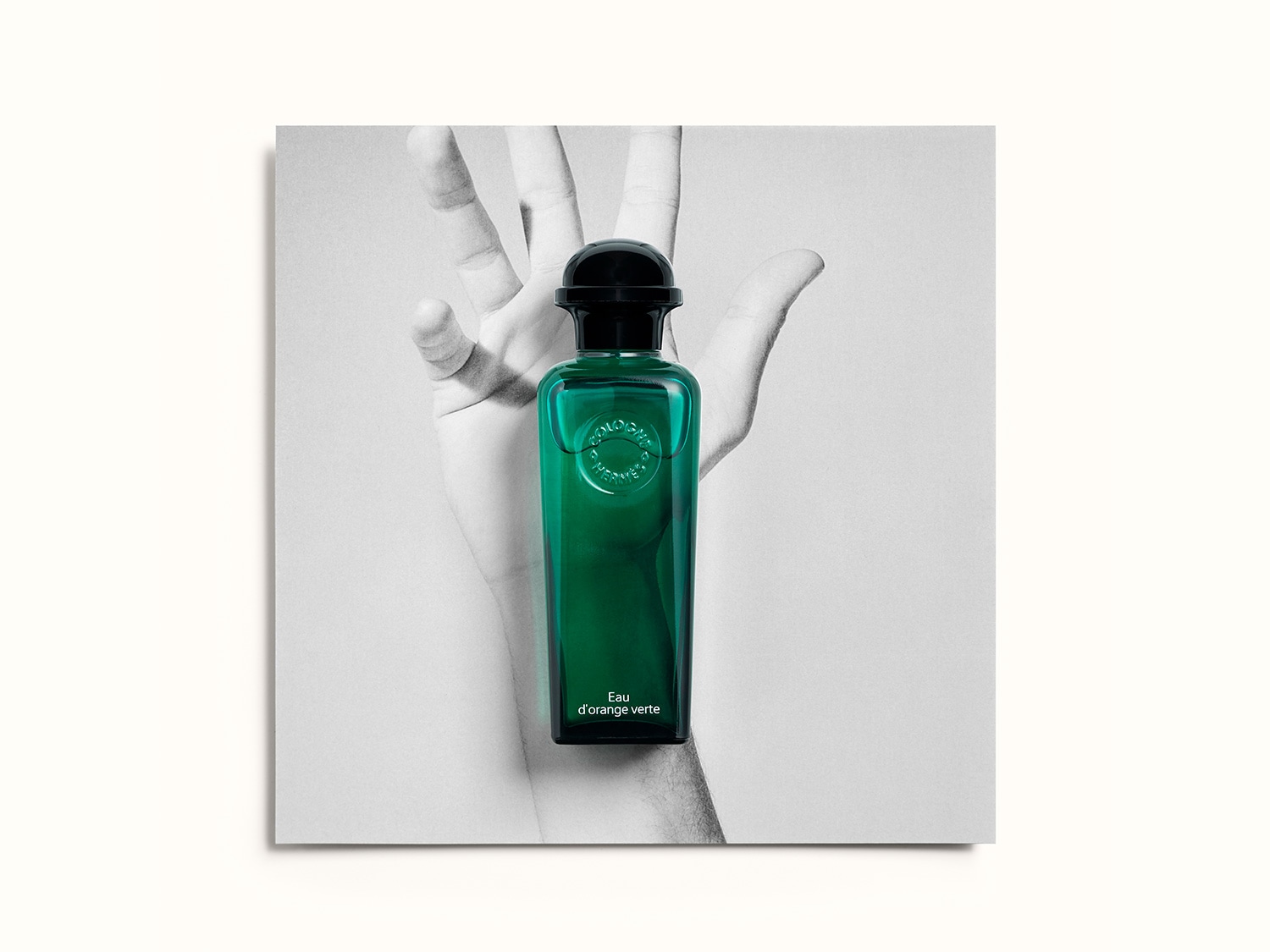 Frasco de perfume verde esmeralda 'Eau d'orange verte' de Hermès, con tapón negro, sostenido por una mano en blanco y negro, evocando una sensación de frescura y sofisticación.