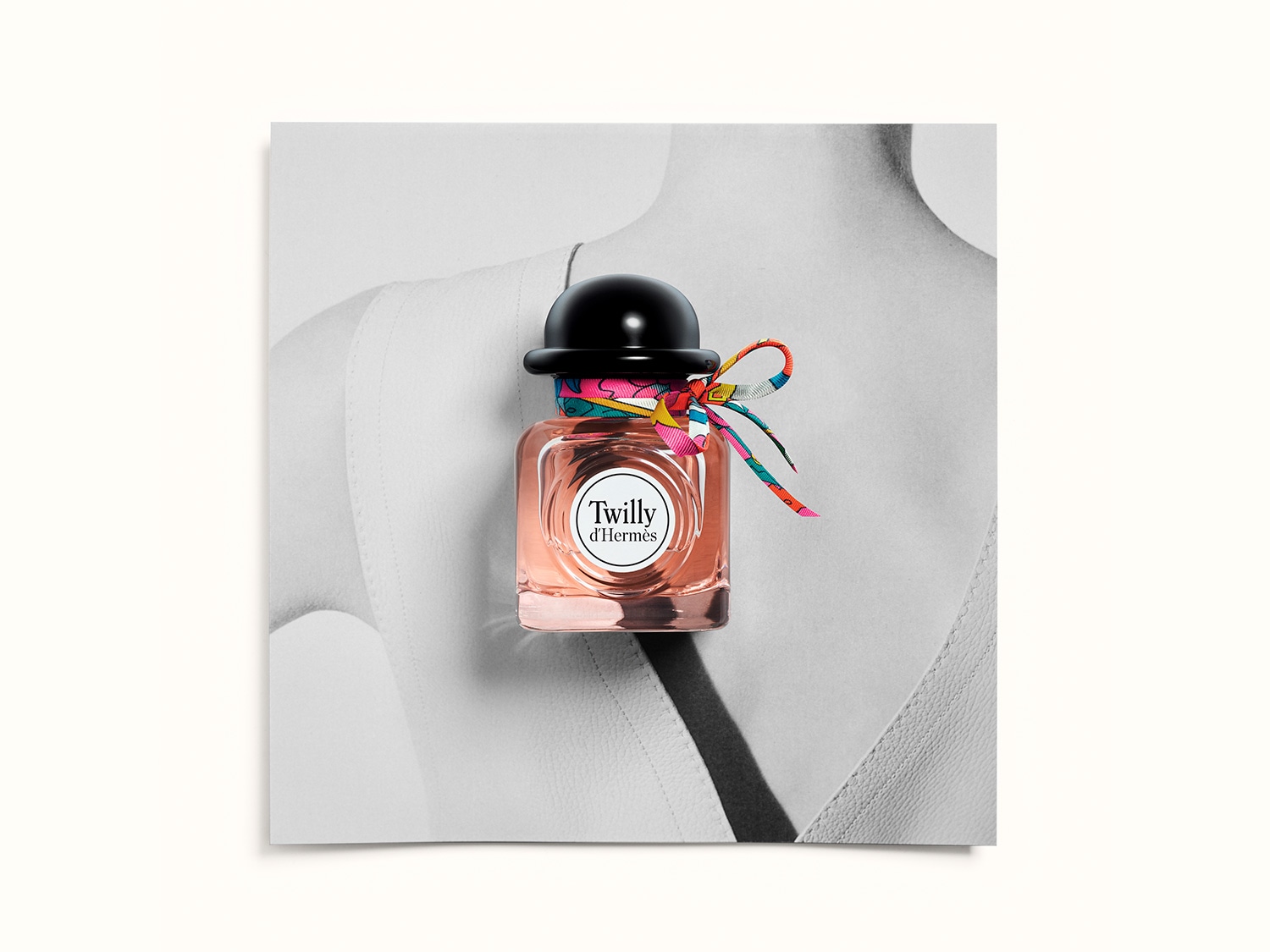 El frasco de perfume Twilly d'Hermès, con su líquido rosado y tapón negro en forma de sombrero, está adornado con una cinta de seda multicolor anudada, sobre un fondo que simula la silueta de una persona.