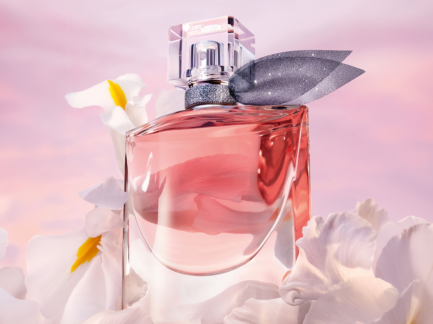 Un frasco de perfume de cristal transparente con líquido rosa, adornado con un lazo brillante y rodeado de pétalos de flores blancas, sobre un fondo rosa claro.