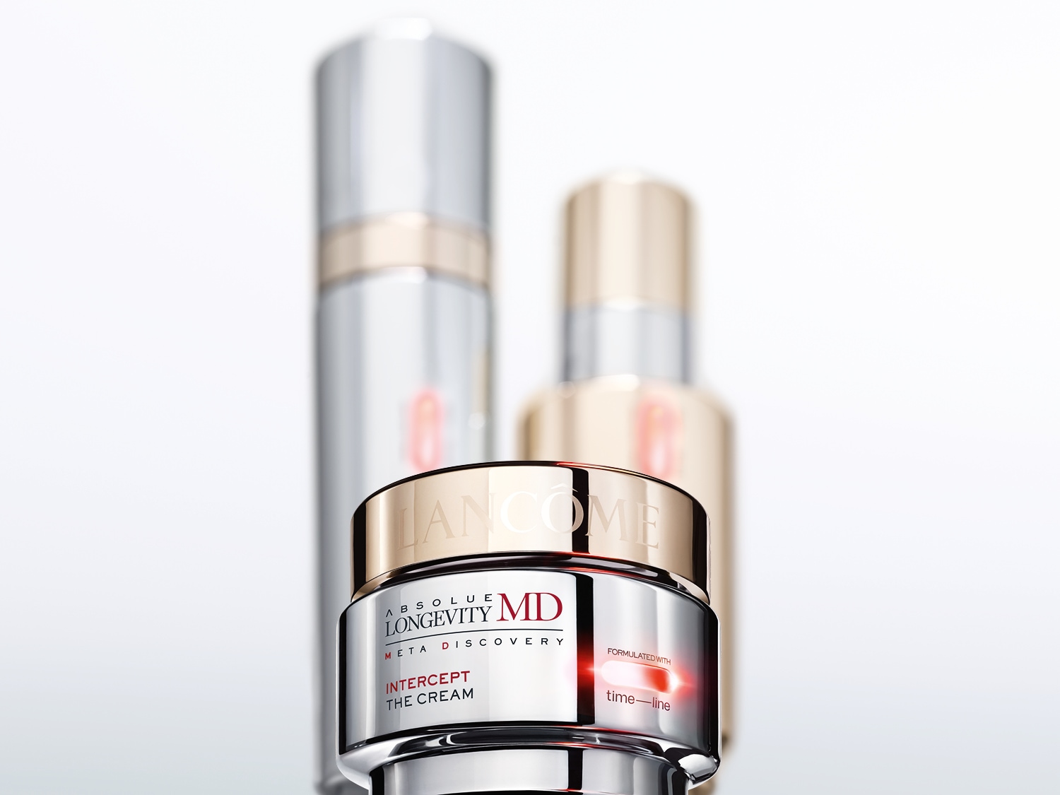 Un primer plano de la crema Lancôme Absolue Longevity MD Intercept The Cream en un envase plateado brillante con tapa dorada, con dos botellas borrosas de Lancôme en el fondo.