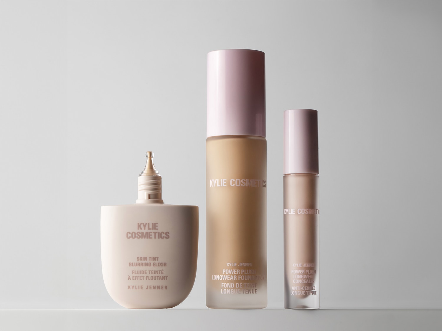 Na zdjęciu znajdują się trzy produkty do makijażu marki Kylie Cosmetics: eliksir do twarzy w beżowym opakowaniu, podkład do twarzy w szklanej butelce z różowym wieczkiem oraz korektor w smukłym opakowaniu z różowym aplikatorem.