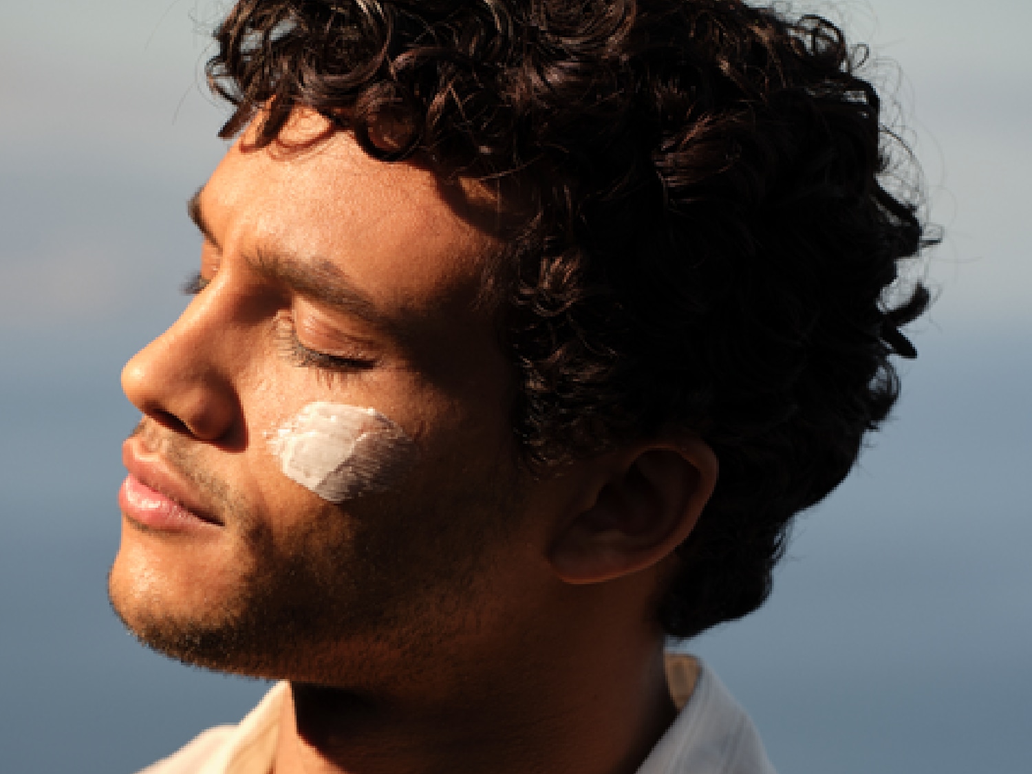 Primer plano del perfil de un hombre moreno con el pelo rizado y una mancha de crema blanca en la mejilla, disfrutando del sol con los ojos cerrados.