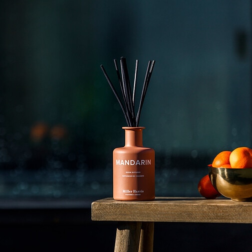 Brązowy dyfuzor zapachowy Miller Harris Mandarin z czarnymi patyczkami na drewnianym stoliku, obok złotej misy z pomarańczami.