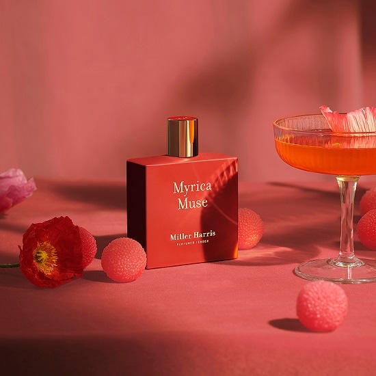 Na tle koralowej scenerii stoi flakon perfum Myrica Muse Miller Harris o kwadratowym kształcie i złotym korku, otoczony kwiatami i kieliszkiem koktajlu.