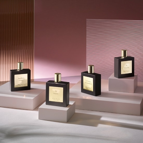 Cztery czarne flakony perfum Miller Harris z złotymi etykietami i korkami, każdy umieszczony na oddzielnym postumencie, prezentowane na tle w odcieniach różu.