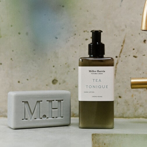 Szare mydło z wytłoczonymi literami M.H. oraz butelka balsamu do rąk Miller Harris Tea Tonique z czarną pompką.