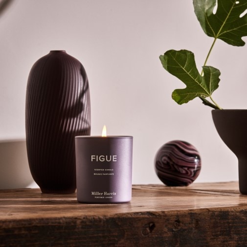 Fioletowa świeca zapachowa Figue marki Miller Harris stoi na drewnianym stole obok wazonów i gałązki figi, tworząc przytulną atmosferę.