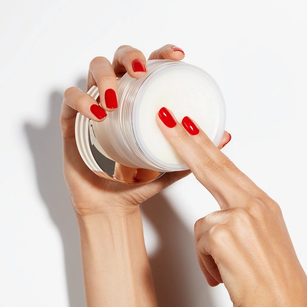 Une main avec des ongles rouge vif plonge un doigt dans un pot transparent rempli d'une crème blanche et épaisse, promettant une hydratation intense.