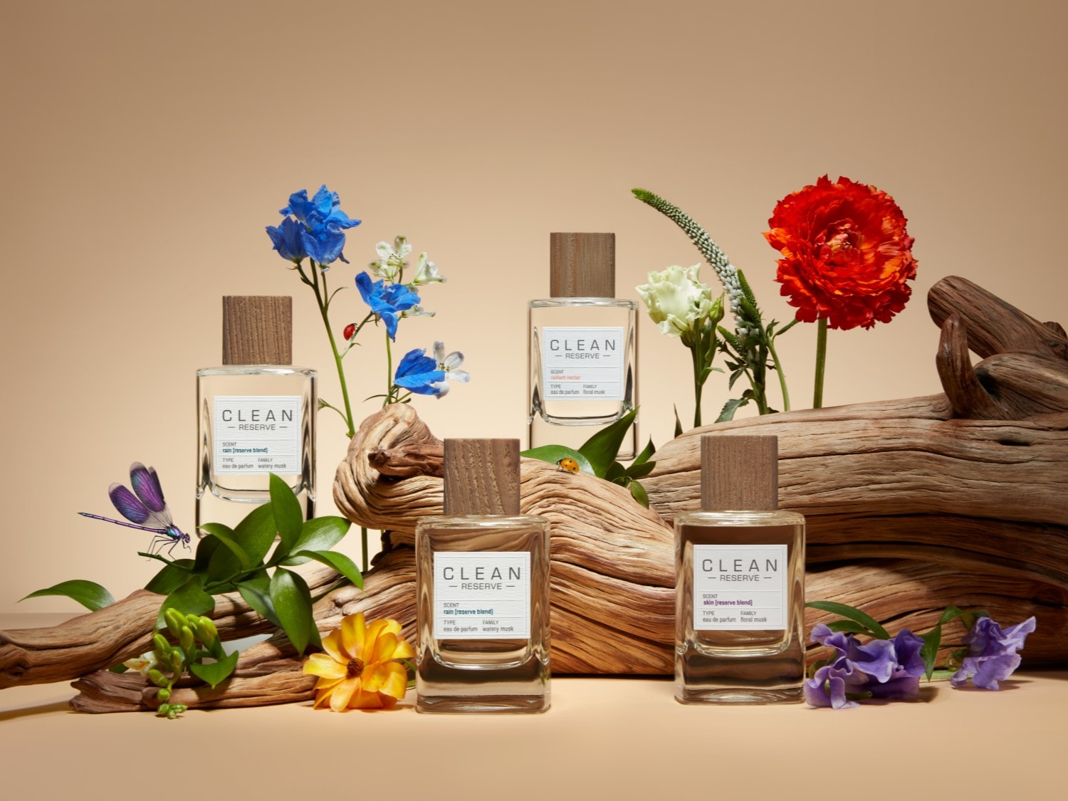 Cztery flakony perfum Clean Reserve z drewnianymi korkami, otoczone kwiatami i gałęziami, tworzą naturalną kompozycję zapachową.