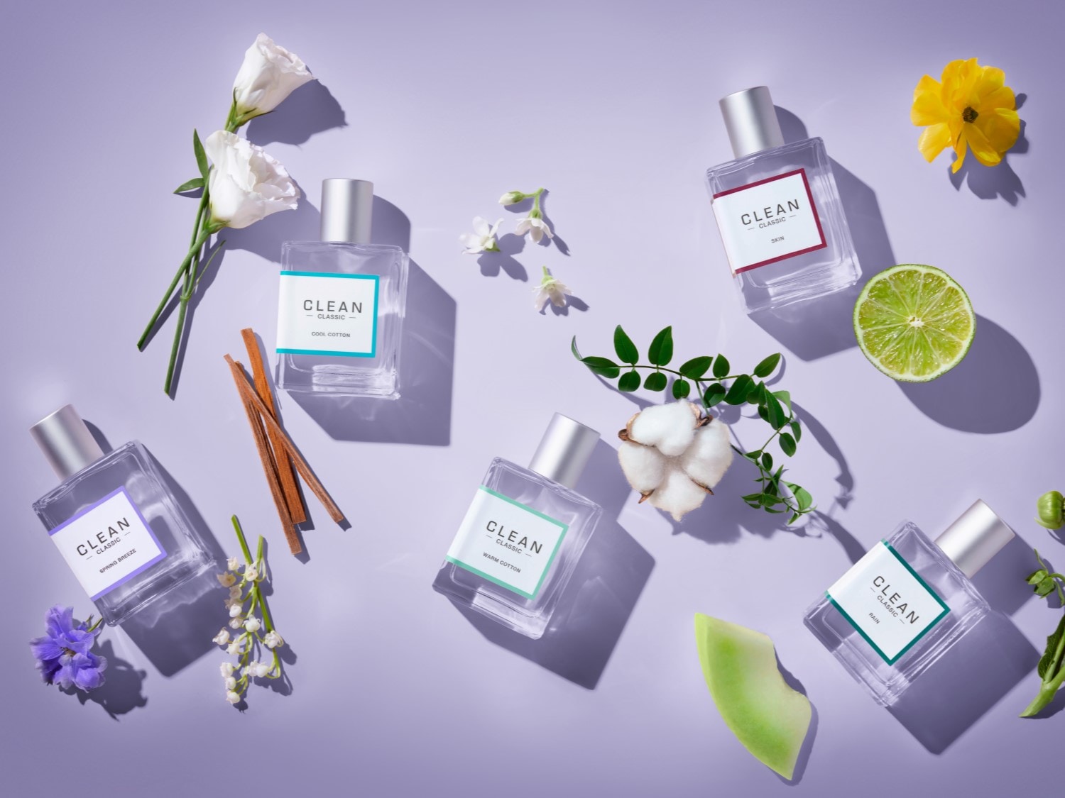 Na fioletowym tle leży sześć flakonów perfum Clean Classic z różnymi zapachami, takimi jak Cool Cotton, Spring Breeze i Rain, otoczonych kwiatami, owocami i przyprawami.