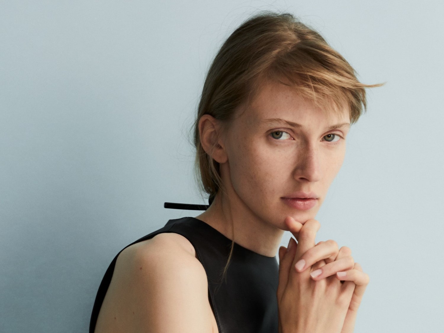 Eine Frau mit hellbraunem, schulterlangem Haar und einem schwarzen Oberteil posiert vor einem hellblauen Hintergrund und hält ihre Hände unter ihr Kinn.