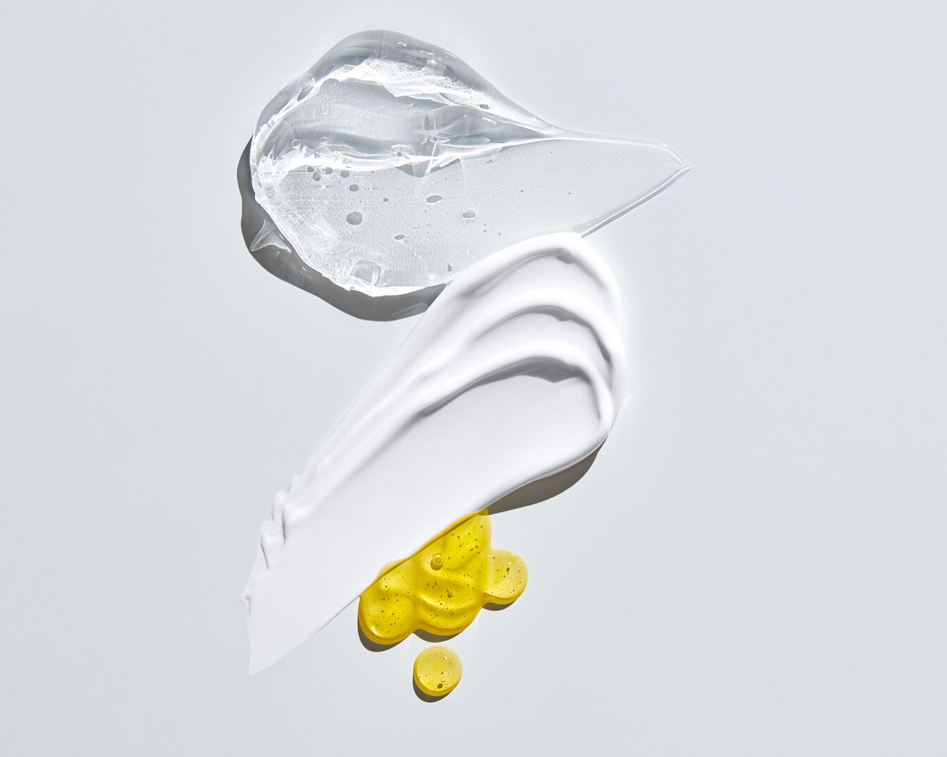 Se ven tres tipos de cosméticos: uno transparente en gel, otro blanco cremoso y un aceite amarillo, todos sobre un fondo blanco.