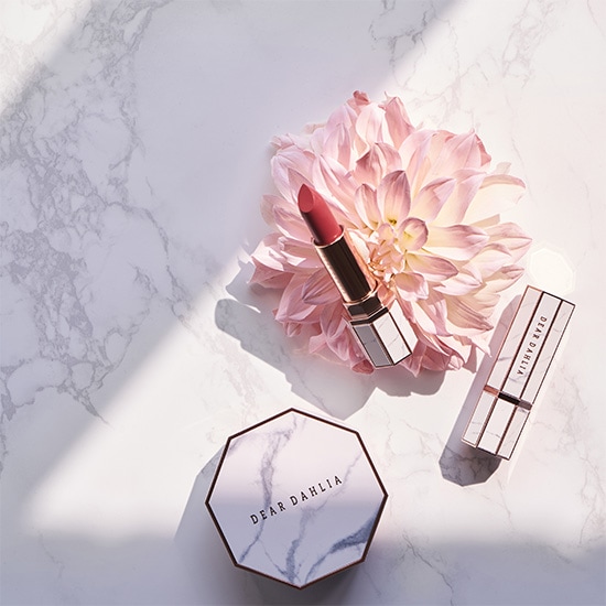 Na marmurowym tle leżą produkty do makijażu Dear Dahlia w odcieniach różu i bieli, obok rozłożystego kwiatu w podobnej kolorystyce.
