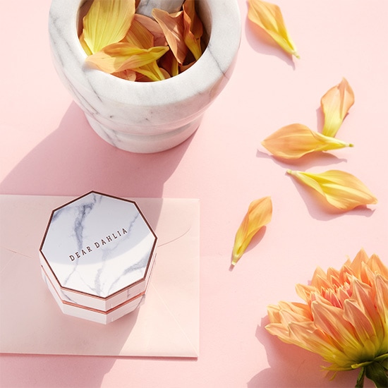 Na różowym tle leży marmurowy moździerz wypełniony płatkami kwiatów, obok znajduje się pudełko Dear Dahlia na różowej kopercie, a całość dopełniają rozsypane płatki i kwiat.