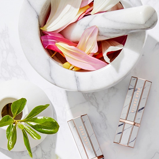 Dwa szminki Dear Dahlia w marmurkowym opakowaniu leżą obok moździerza wypełnionego płatkami kwiatów i małej rośliny doniczkowej.