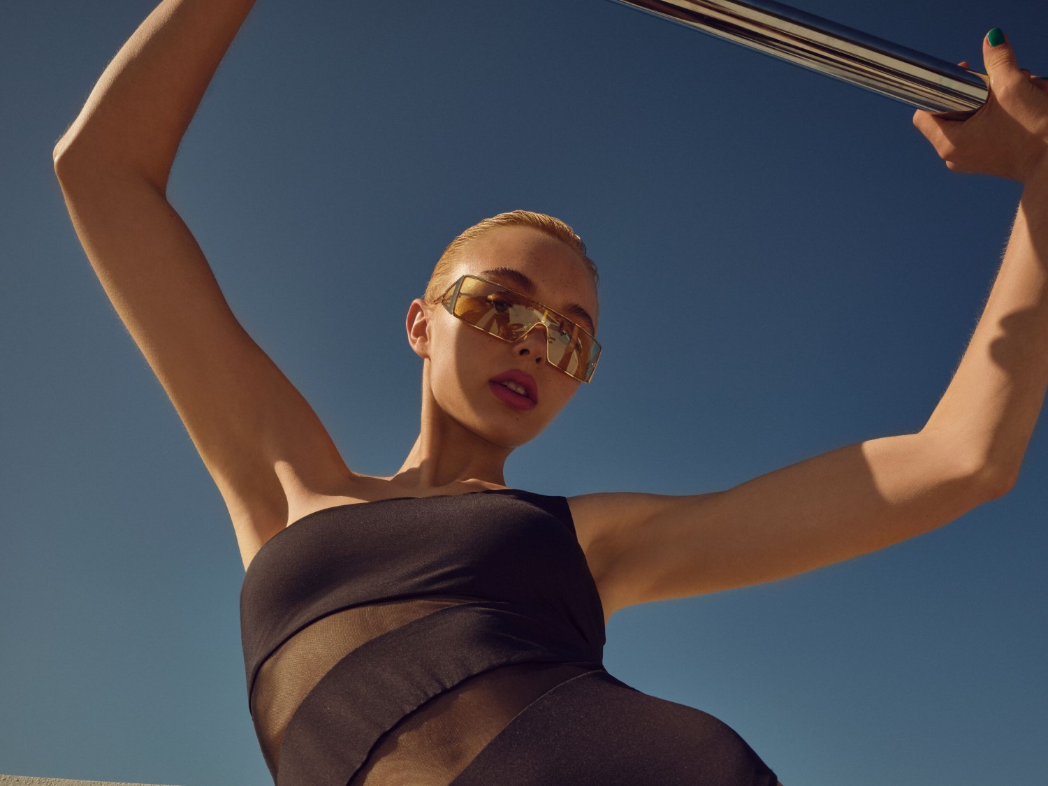 Eine Frau mit blonden, zurückgekämmten Haaren trägt einen schwarzen Badeanzug mit transparenten Einsätzen und eine goldfarbene Sonnenbrille vor einem strahlend blauen Himmel.