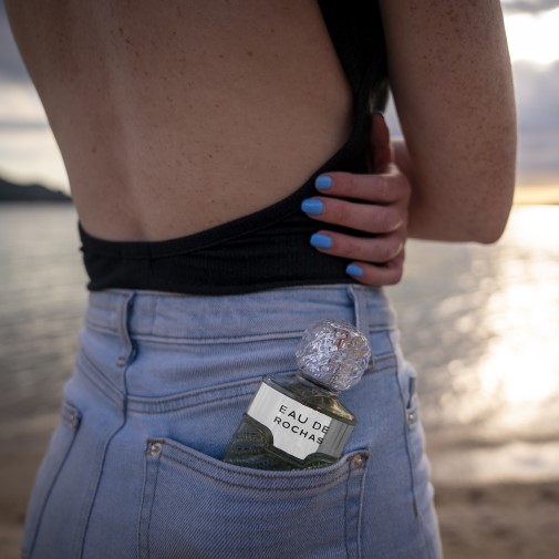 Butelka perfum Eau de Rochas schowana w tylnej kieszeni dżinsów kobiety na tle plaży o zachodzie słońca.