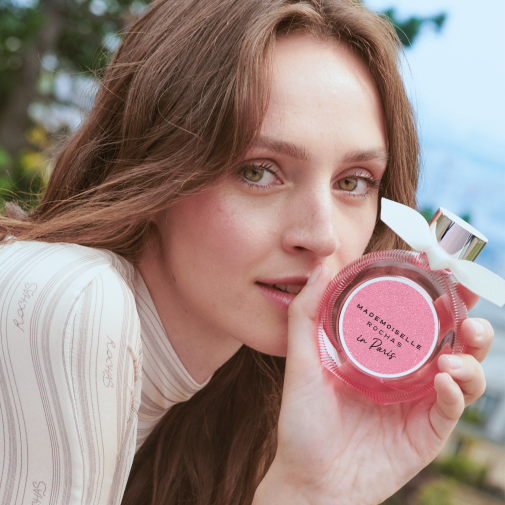 Młoda kobieta prezentuje okrągły flakon perfum Mademoiselle Rochas In Paris w różowym opakowaniu z białą kokardką, emanujący świeżością i paryskim szykiem.