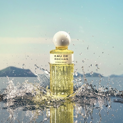 Butelka perfum Eau de Rochas Citron Soleil z żółtym płynem i białą nakrętką stoi na tle morza i nieba, otoczona rozpryskującą się wodą, co sugeruje świeżość i lekkość zapachu.