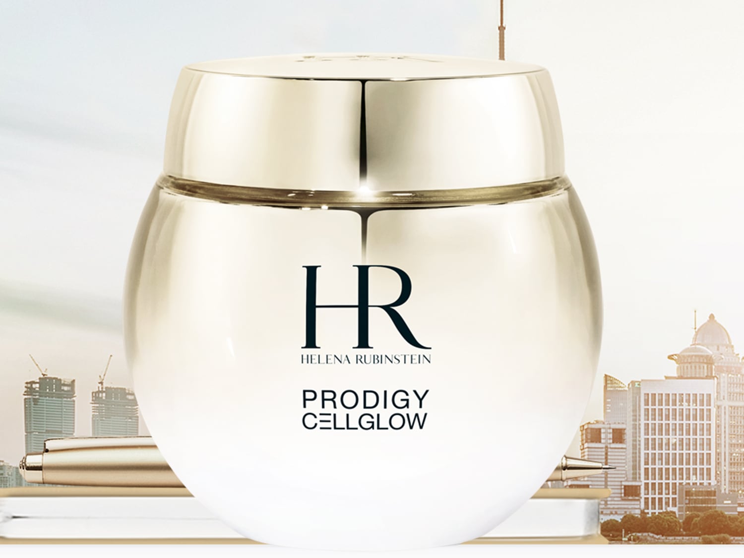 Srebrny słoiczek kremu Helena Rubinstein Prodigy Cellglow na tle panoramy miasta, obok leży złote pióro.