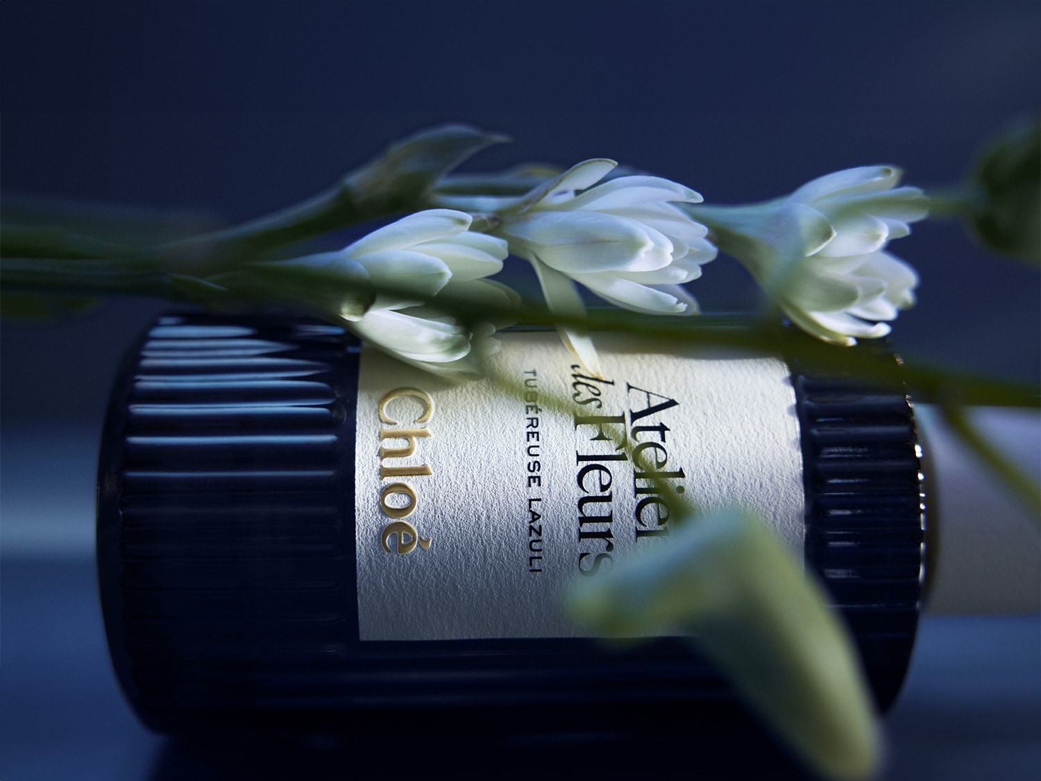 Butelka perfum Chloé Atelier des Fleurs Tubéreuse Lazuli leży na boku, ozdobiona białymi kwiatami, prezentując elegancję i kwiatowy aromat.