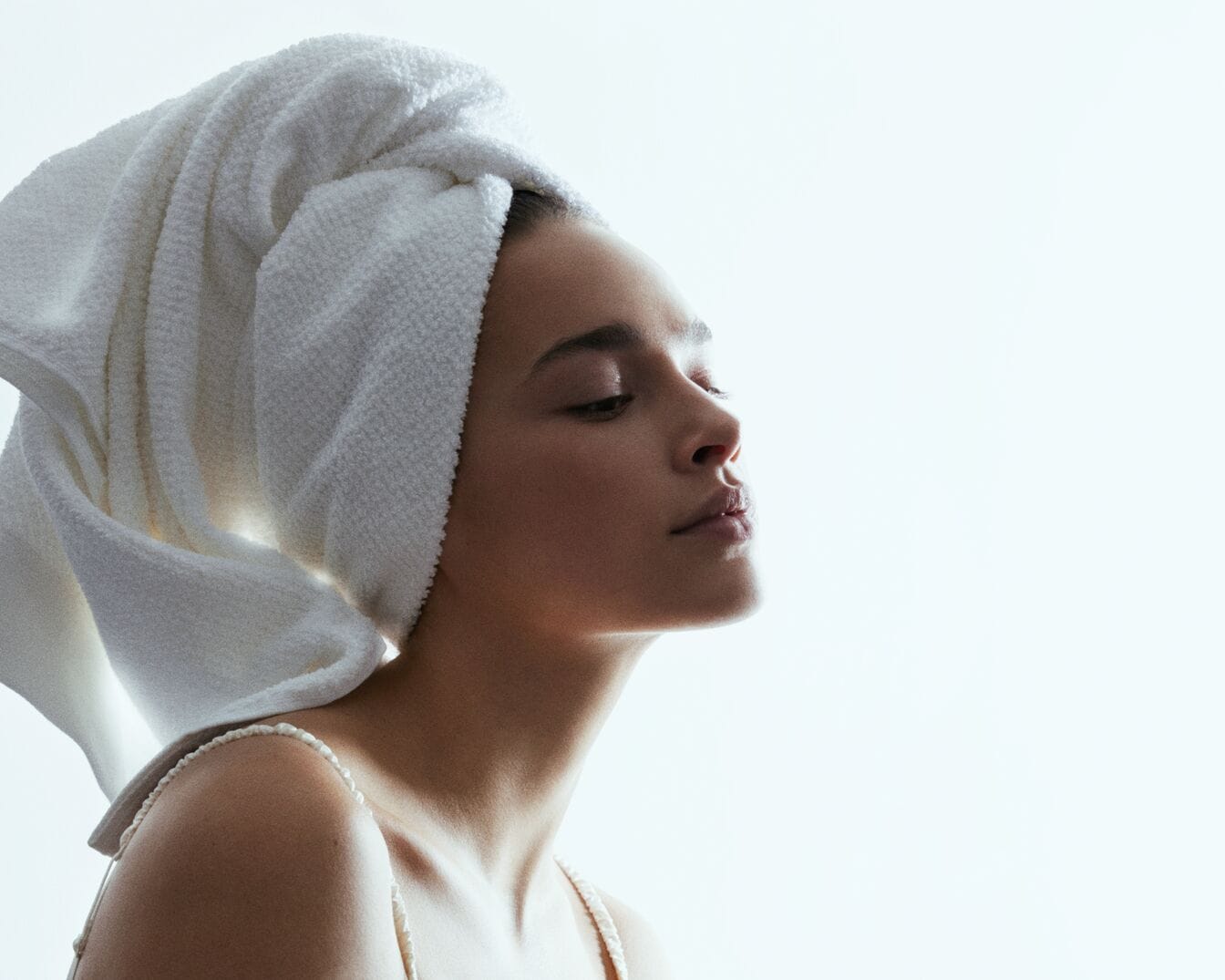 Una mujer con una toalla blanca en la cabeza, con los ojos cerrados y la piel suave, disfrutando de un momento de relajación y cuidado personal.