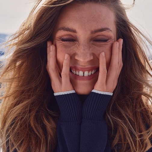 Nahaufnahme einer strahlenden Frau mit geschlossenen Augen, die ihr Gesicht mit den Händen umrahmt und ein breites Lächeln zeigt.