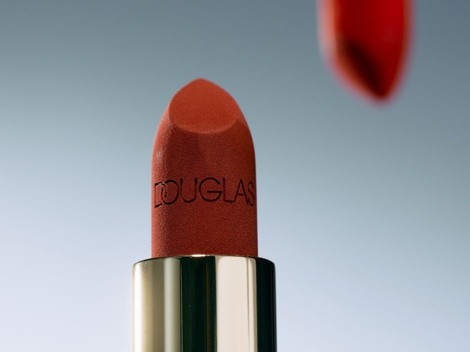 Een close-up van een Douglas lipstick in een warme, roestkleurige tint, met de merknaam subtiel in de lipstick gegraveerd, gevat in een elegante gouden huls.