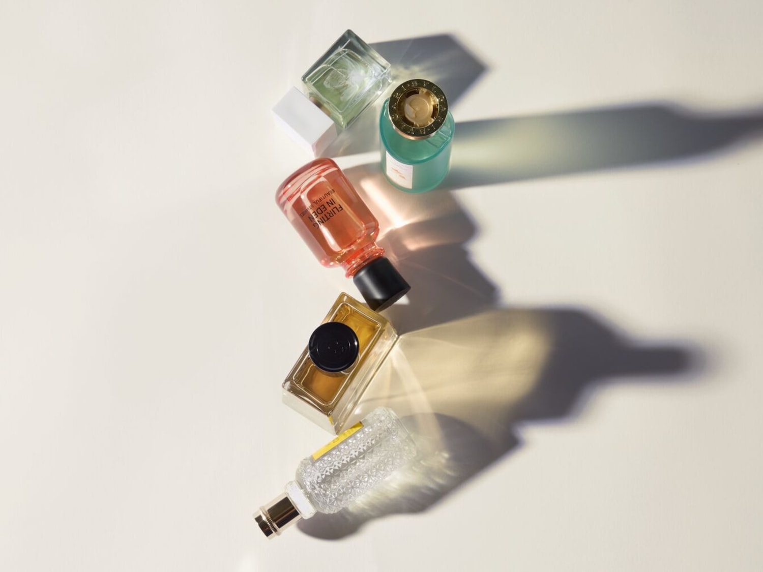 Een bovenaanzicht van een verzameling parfumflesjes in verschillende kleuren en vormen, waaronder roze, groen, goud en helder, die een reeks schaduwen werpen op een heldere achtergrond.