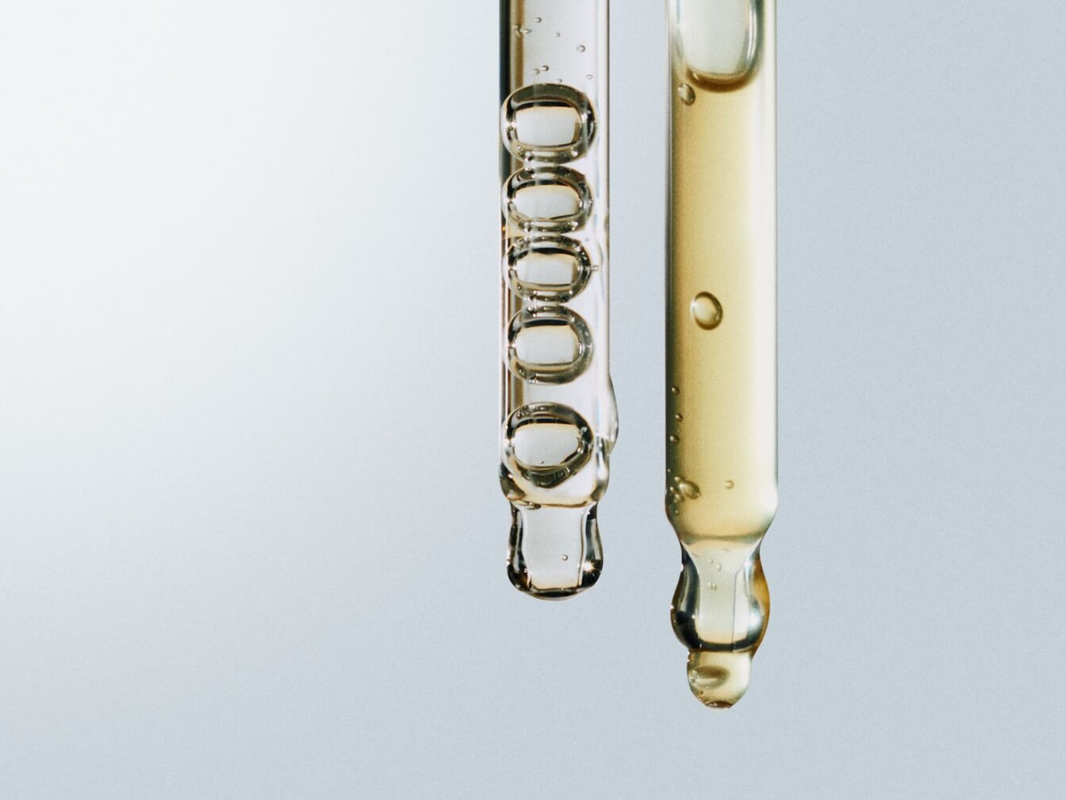 Twee glazen pipetten, één gevuld met heldere vloeistof en de andere met een goudkleurige olie, klaar voor gebruik in je huidverzorgingsroutine.