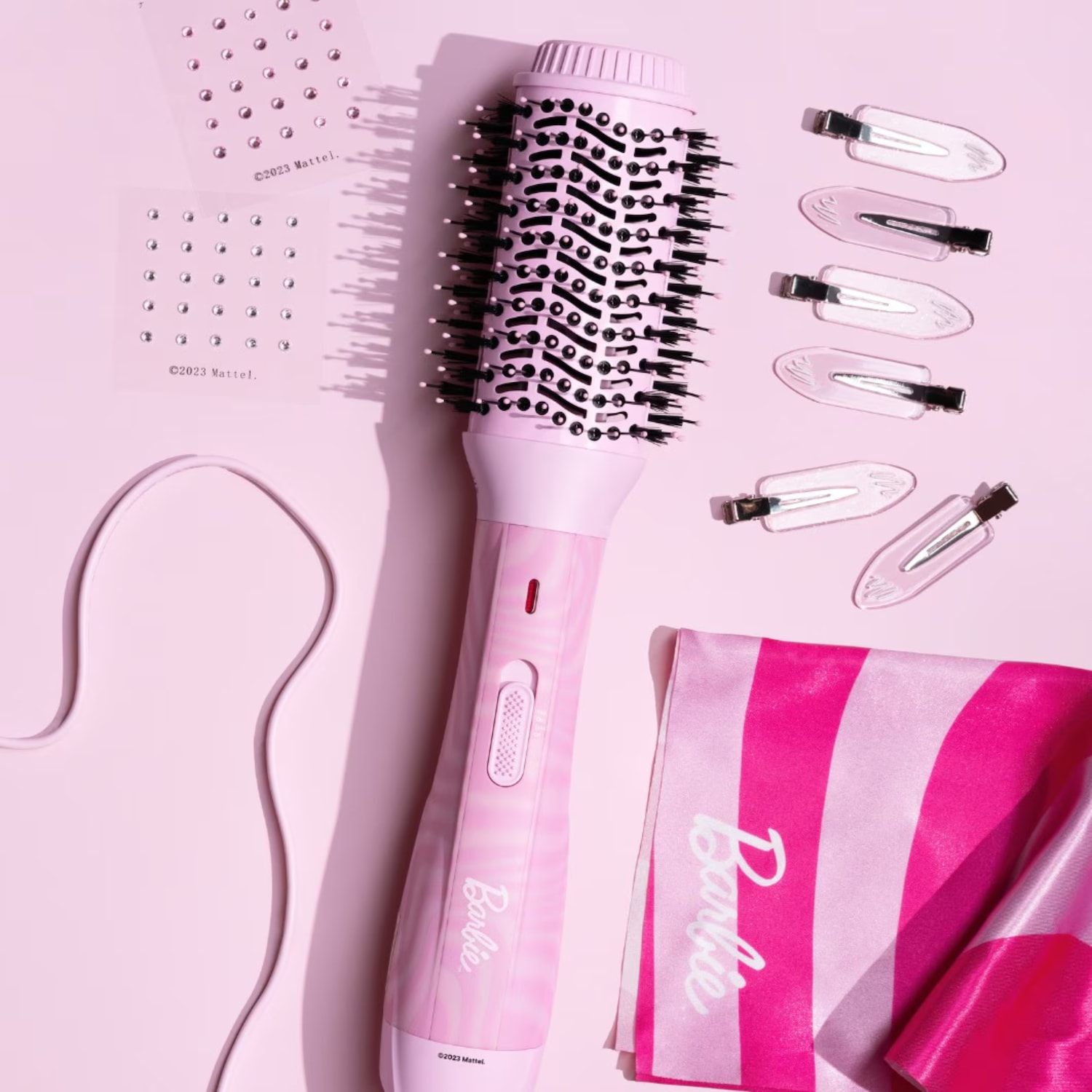 De roze Barbie föhnborstel met zwarte borstelharen, samen met transparante haarclips en twee vellen met zilveren strass-steentjes, ligt op een lichtroze achtergrond, met een roze en wit gestreepte handdoek met het Barbie-logo in wit.