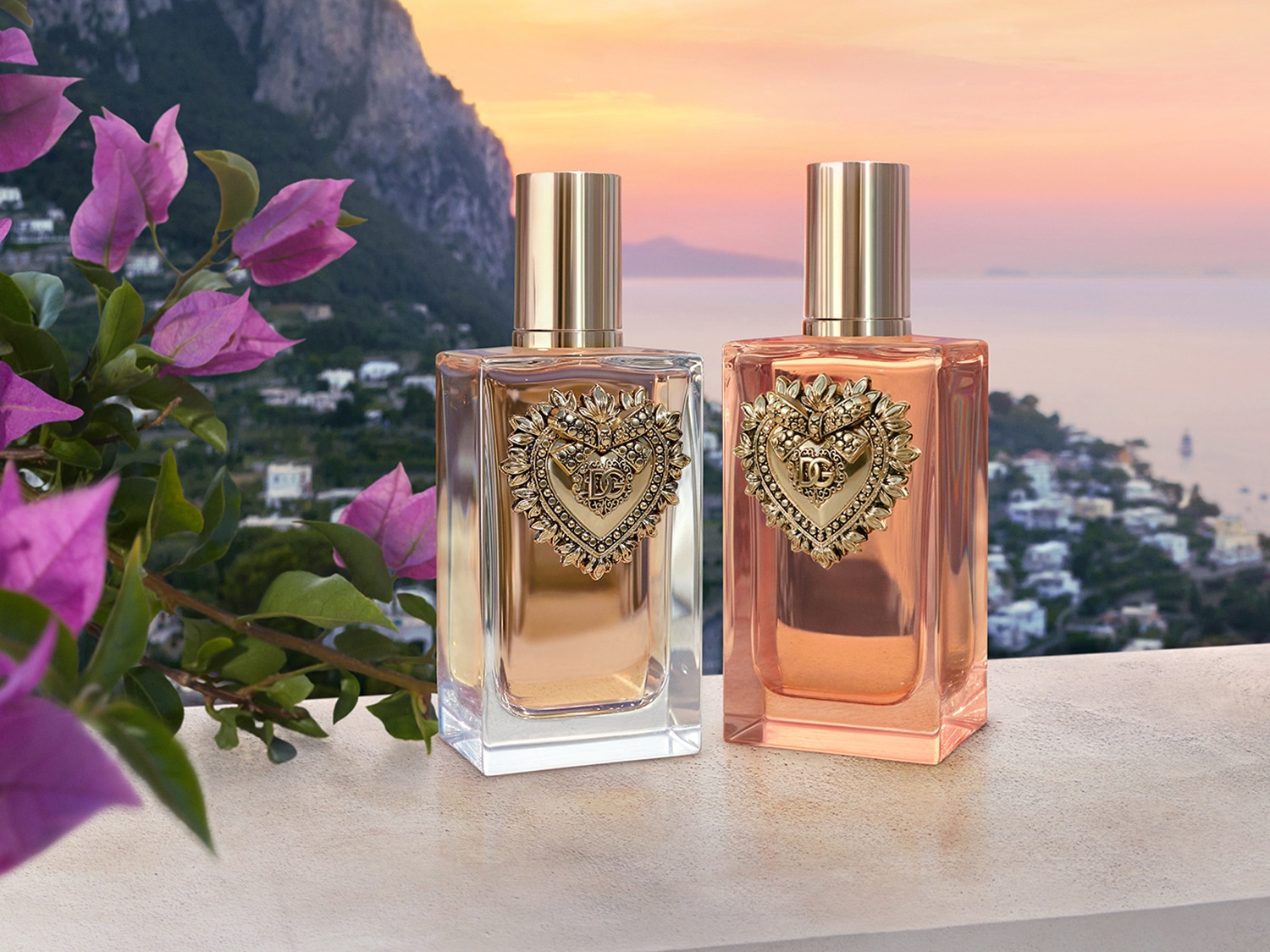 Dwa flakony perfum Dolce&Gabbana Devotion, jeden w odcieniu złotym, drugi różowym, oba z dekoracyjnym złotym sercem na tle malowniczego krajobrazu.