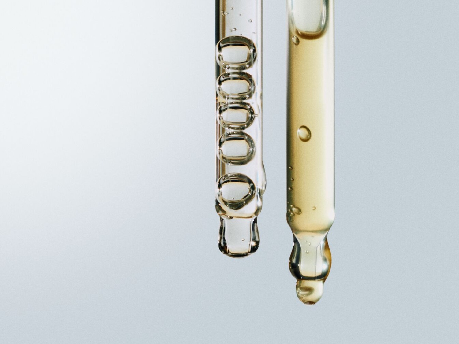 Twee glazen pipetten, één gevuld met heldere vloeistof en de andere met een goudkleurige vloeistof, beide met een druppel aan de punt.
