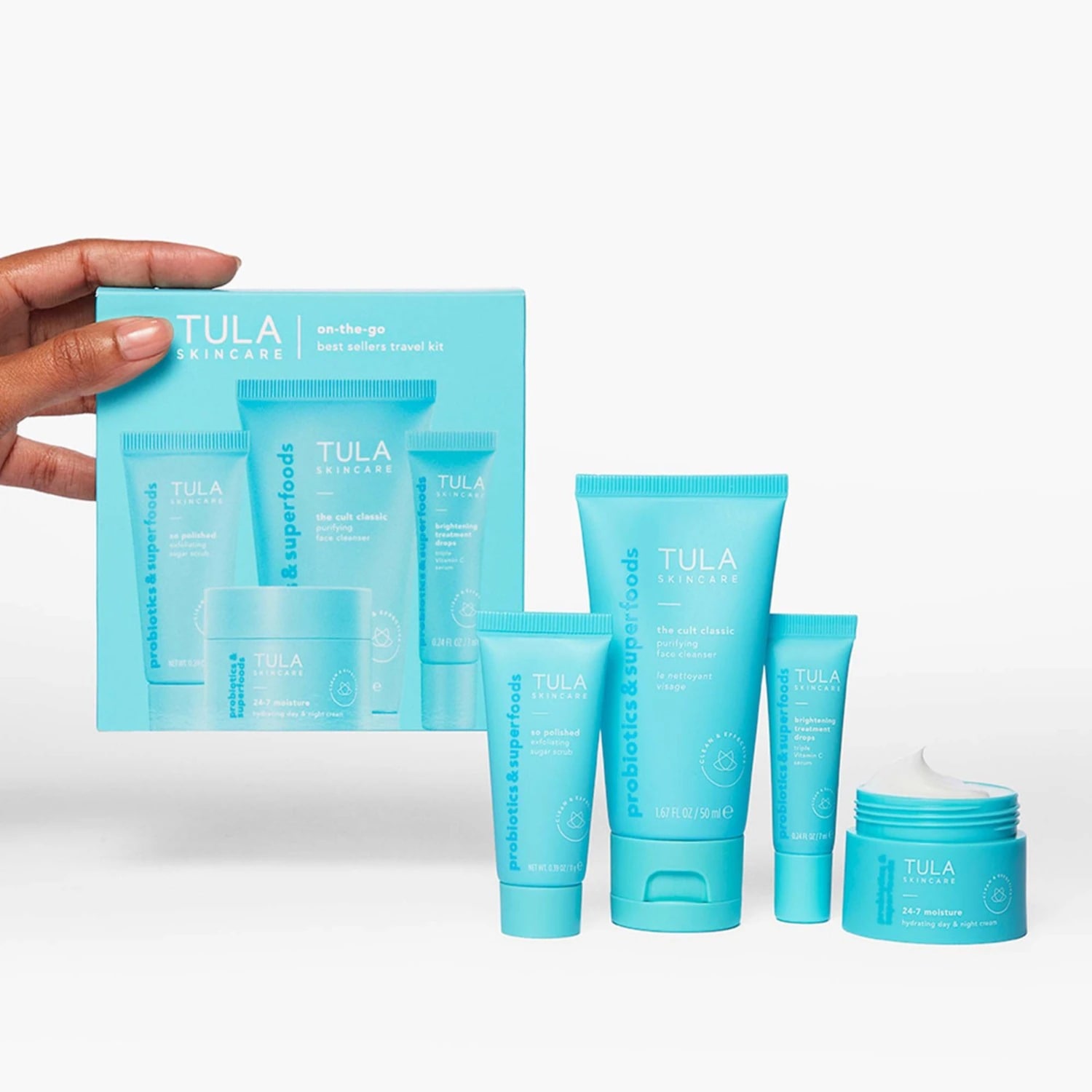 Une main tient une boîte bleu clair "TULA Skincare on-the-go best sellers travel kit" contenant quatre produits de soin, tandis que quatre autres produits sont disposés devant la boîte, dont un pot de crème hydratante ouvert.