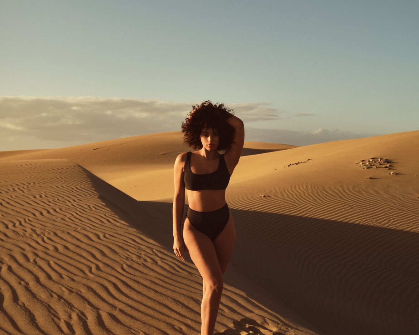 Eine Frau mit lockigem Haar und dunklem Bikini steht in einer Sandwüste unter einem hellblauen Himmel.
