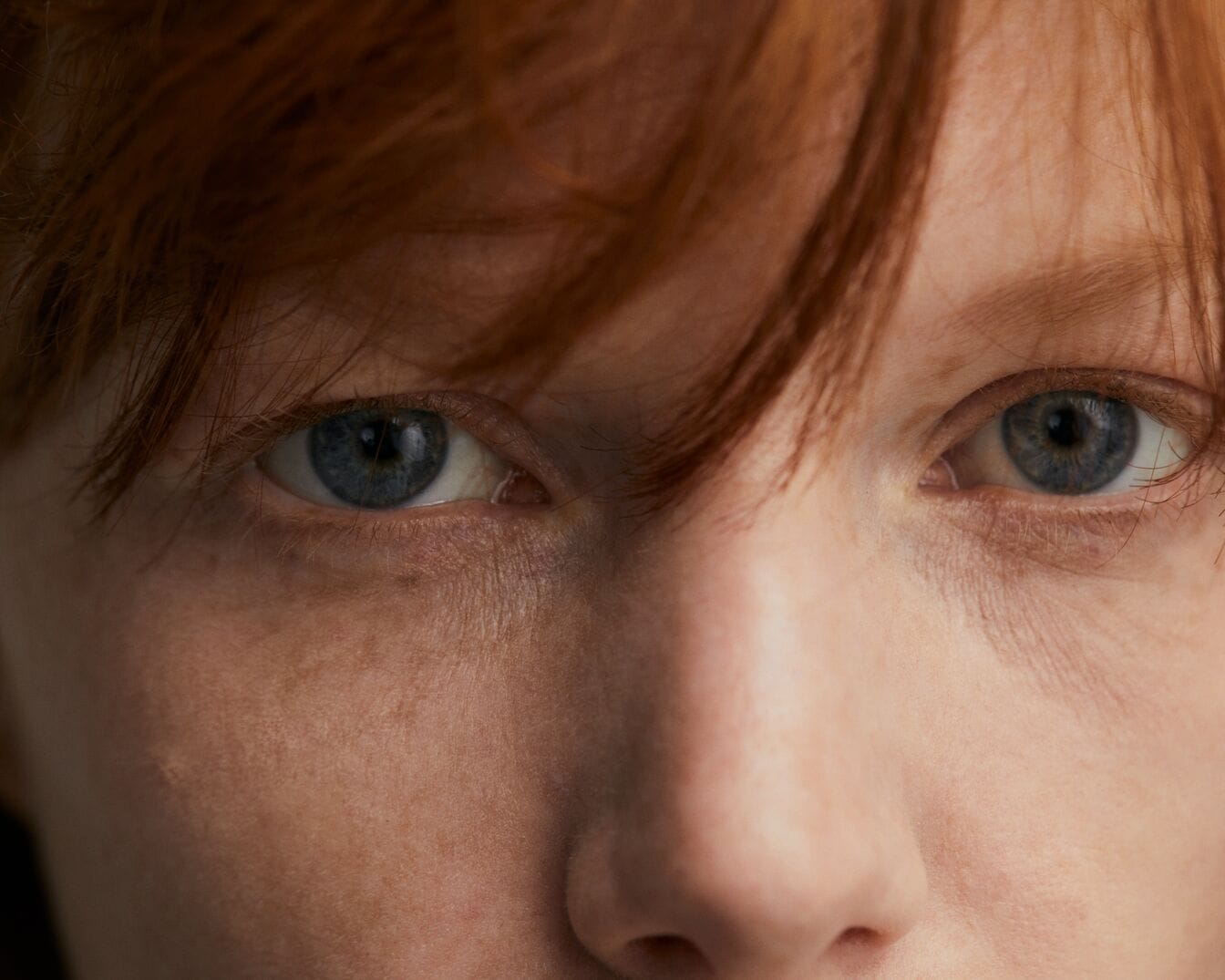 Nahaufnahme eines Frauengesichts mit hellblauen Augen und roten Haaren, die Stirn bedecken.