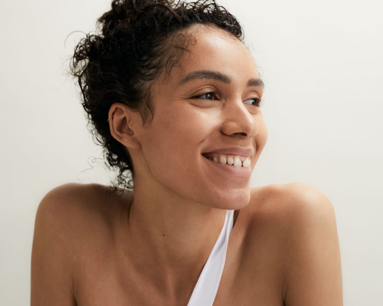 Nahaufnahme einer strahlenden Frau mit lockigem, dunklem Haar, die ein weißes Trägeroberteil trägt und mit einem herzlichen Lächeln zur Seite blickt.