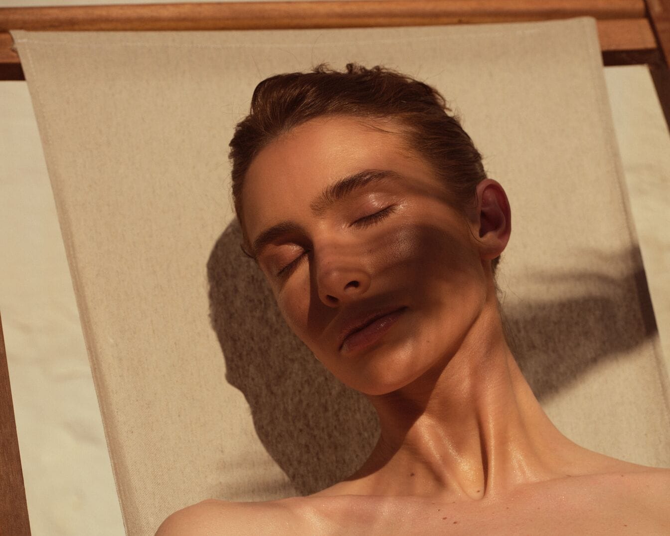 Eine Frau liegt mit geschlossenen Augen auf einem Liegestuhl, ihr Gesicht ist dem Sonnenlicht zugewandt, das Schatten auf ihr Gesicht wirft.