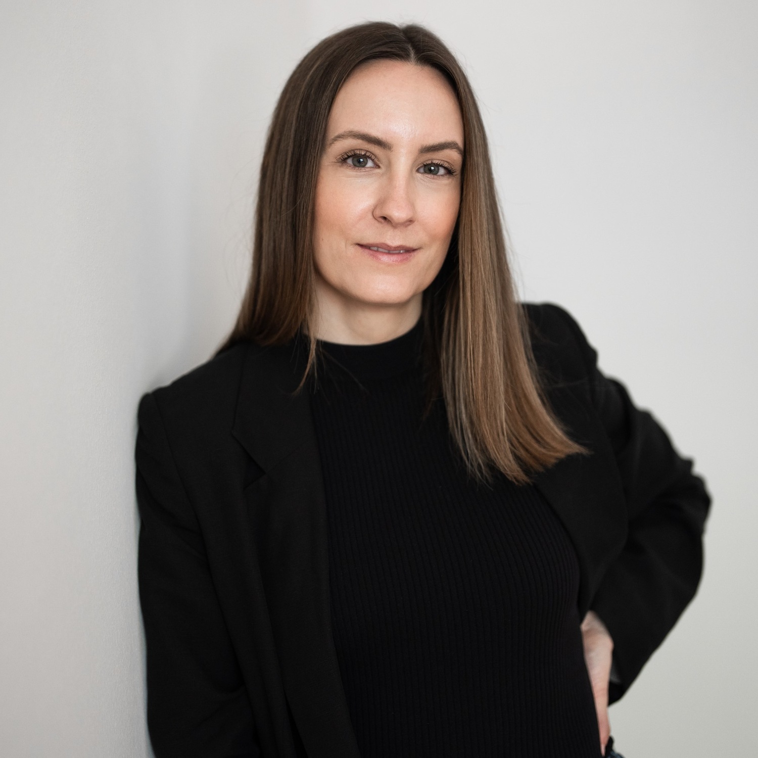 Eine Frau mit langen braunen Haaren, gekleidet in einen schwarzen Blazer und ein schwarzes Oberteil, lehnt lächelnd an einer weißen Wand.