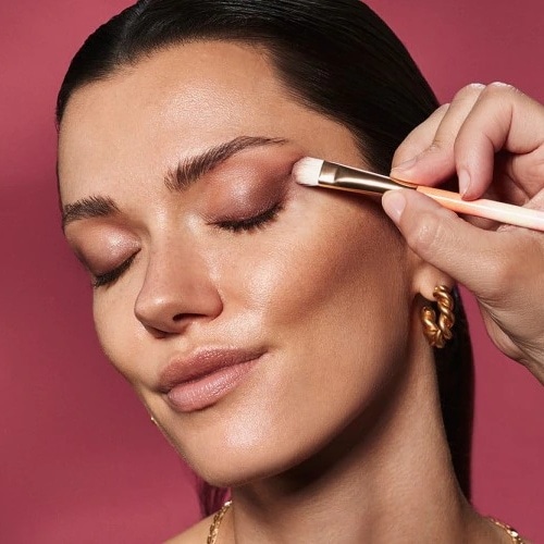 Con los ojos cerrados, una mujer se aplica sombra de ojos rosa brillante con una brocha de mango dorado para un look glamuroso.