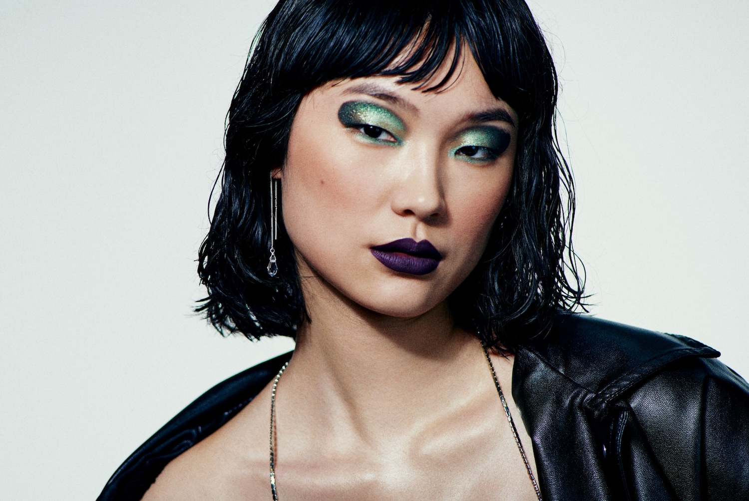A képen egy nő látható, aki fekete bőrdzsekit visel, sminkje pedig zöld szemhéjfestékből és sötétlila rúzsbol áll.