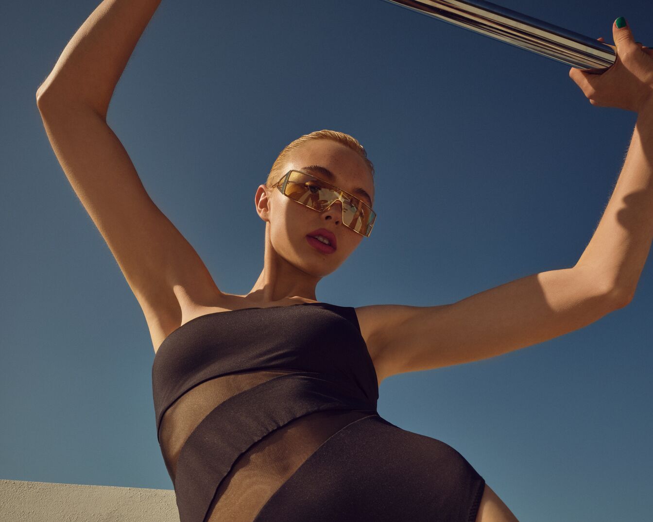 Una mujer con gafas de sol doradas y un traje de baño negro posa con un cielo azul despejado de fondo.