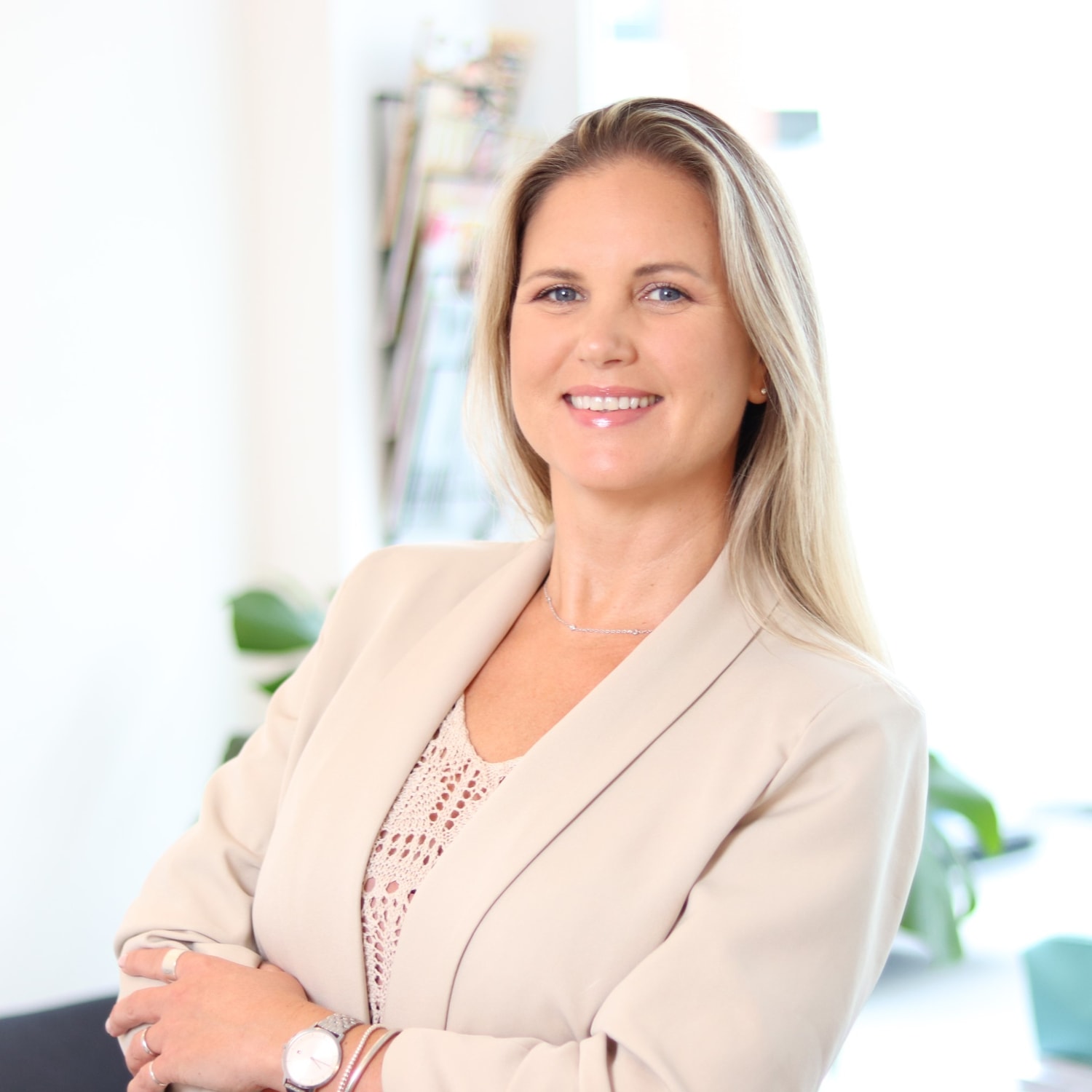 Eine blonde Frau mit einem beigen Blazer lächelt mit verschränkten Armen und trägt eine silberne Uhr.