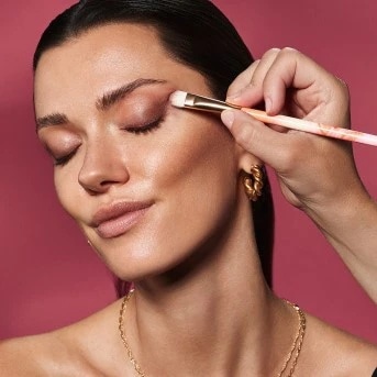 Una mujer se está aplicando sombra de ojos rosa con un pincel para crear un look de maquillaje suave y elegante.