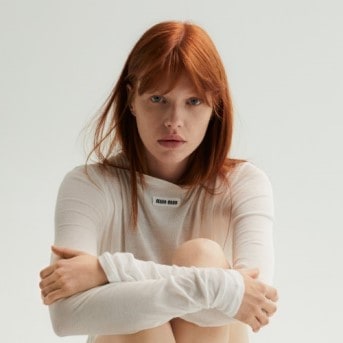 Eine Frau mit roten Haaren und hellem Teint umarmt ihre Knie, während sie ein langärmeliges Oberteil mit einem kleinen Logo trägt.