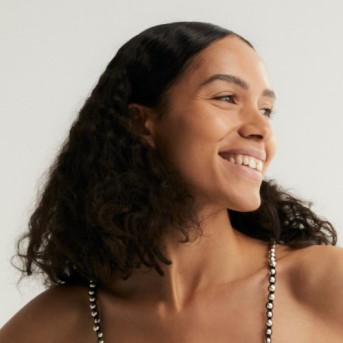 Nahaufnahme einer lächelnden Frau mit dunklem, gewelltem Haar und einem trägerlosen Oberteil, das mit silbernen Perlen verziert ist.