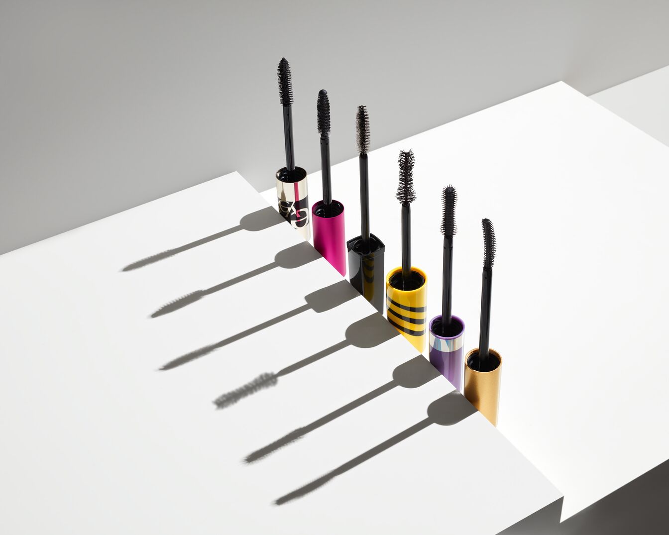 Seis máscaras de pestañas de diferentes colores, incluyendo dorado, fucsia, amarillo y morado, se alinean sobre una superficie blanca, cada una con su aplicador negro sobresaliendo.