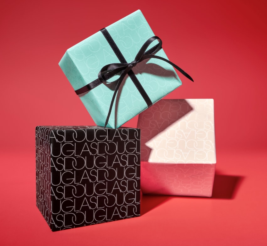 Na czerwonym tle piramida z trzech prezentów Douglas: turkusowy z czarną wstążką na górze, pod nim czarny z białym napisem Douglas i różowy z jasnoróżowym napisem Douglas.